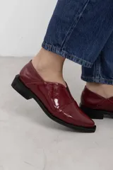 Mocasines de charol color bordó, de diseño vanguardista con corte asimétrico en el empeine, abertura en V, punta fina y suela plana con pequeño tacón.