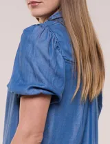 Camisa de lyocell color azul con efecto denim, cuello camisero, cierre frontal con botones y detalles de bordado oscuro a lo largo de la botonera. Presenta mangas cortas abullonadas con puños elásticos y terminación con volados.