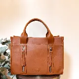 Cartera de cuero color tostado, estructurada, con doble asa de mano, correa desmontable y dos bolsillos frontales verticales con cierre. Presenta detalles de costuras pespunteadas y el logo de la marca grabado en la parte inferior.