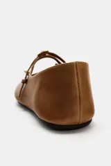Zapato tipo bailarina de piel color marrón tostado, con tira delantera ajustable, aplique metálico y punta redonda.