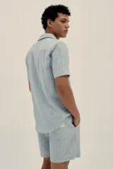 Camisa celeste a rayas verticales, de manga corta, con cuello tipo camp-collar, botones de madera y parche con logo.