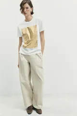 Camiseta de algodón con cuello redondo y manga corta. Presenta un diseño de estampación metalizada en color dorado en la parte frontal con detalles de texto.