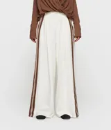 Pantalón ancho de corte oversize, color blanco, con cintura alta ajustable con cordón. Presenta pliegue frontal profundo y cintas a rayas marrones en las costuras laterales.