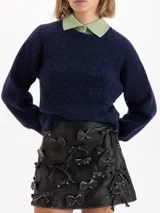 Mini falda de cuero ovino negro con efecto brush off, decorada con múltiples apliques de moños de cuero fijados con remaches metálicos. Posee bolsillos ojal en la parte trasera y forrería de acetato.