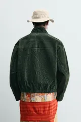 Cazadora regular fit de piel con acabado en ante color verde oscuro, de la colección especial Gabriel Azevedo x ZARA. Cuenta con cuello subido, manga larga, bolsillos de vivo en la cadera, bolsillo interior y bajo acabado con elástico. Cierre frontal con cremallera oculta por solapa con botones a presión.