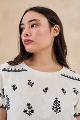 Remera de algodón color blanco con bordados florales y geométricos en color negro. Presenta cuello redondo, mangas cortas y un diseño con franjas horizontales bordadas.