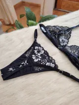 Conjunto de lencería compuesto por un corpiño triangular y una bombacha tipo tanga, confeccionados en encaje negro con detalles florales en hilo plateado.