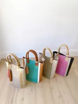 Bolso matero color beige con textura tipo piel de cocodrilo y detalles dorados. Tiene doble asa de mano y un dije decorativo con flecos rosas y marrones.