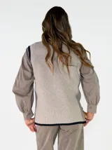 Chaleco de punto en color crema con detalles de ribetes en color marrón oscuro en el cuello, sisas y ruedo. Presenta un diseño clásico con cuello redondo y un pequeño aplique decorativo en el lateral inferior.