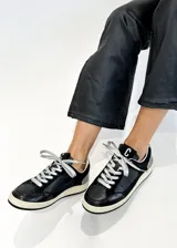 Zapatillas urbanas negras con detalles en cuero, gamuza y charol. Presentan cordones plateados, suela color crema y logo "C" en el lateral.