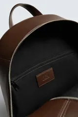 Mochila lisa de color marrón oscuro con acabado mate. Cuenta con un compartimento principal con cierre de cremallera, un bolsillo interior con cremallera y un bolsillo frontal exterior también con cremallera. La parte posterior es acolchada e incluye una banda de sujeción para equipaje. Dispone de un asa de mano superior y dos asas de hombro ajustables.