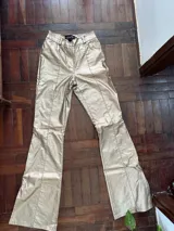 Pantalón de tiro alto confeccionado en eco cuero con acabado metalizado dorado. Presenta un corte recto con aberturas laterales con cierre en los tobillos y costuras frontales decorativas.