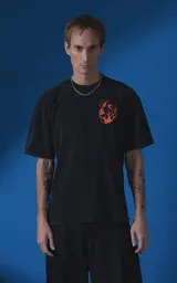 Remera de manga corta con cuello redondo y calce oversize, color negro con efecto nevado. Presenta una estampa frontal en color naranja con diseño gráfico de un rostro rodeado de llamas y texto tipográfico.