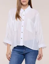 Camisa blanca de silueta holgada y textura liviana, con cuello camisero clásico, abotonadura frontal con botones en contraste y doble bolsillo de parche en el frente. Cuenta con mangas de corte dolman de largo tres cuartos con puño amplio y ruedo recto con aberturas laterales.
