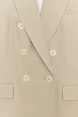 Blazer de mujer color beige claro, de corte largo y cruzado con doble botonadura. Presenta cuello solapa y bolsillos delanteros de ojal.