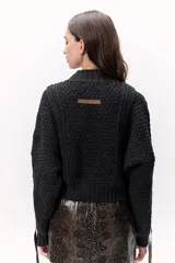 Sweater de lana merino tejido en punto grueso, color gris oscuro, con cuello alto. Presenta un diseño calado con tientos de cuero marrón insertados a lo largo de la prenda, simulando un lazo o detalle orgánico.