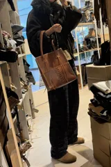 Bolso tipo tote de cuero vacuno color suela, confeccionado artesanalmente con costuras decorativas visibles en los bordes y en el centro. Cuenta con doble asa larga para llevar al hombro y detalles de tiras colgantes en la unión de las asas.