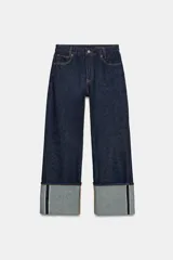 Jeans de tiro bajo y pernera ancha, confeccionados en denim azul oscuro con pespuntes en contraste. Cuentan con un diseño de cinco bolsillos, cierre frontal con cremallera y botón metálico, y destacan por su amplio doblando en el bajo que deja ver el revés de la tela con detalles de costura selvedge.