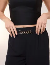 Pantalón negro de calce relajado y caída fluida, confeccionado en poliéster con textura de crepé. Tiene cintura alta elastizada estilo pull on con un aplique de cadena metálica decorativa, y pierna de corte ancho tipo wide leg corto por arriba del tobillo.