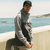Campera canguro Maui and Sons de felpa con cierre completo y capucha ajustable con cordón. Presenta un diseño bicolor en tonos gris claro y gris oscuro, separado por una franja horizontal con patrón de olas y detalles en turquesa. Cuenta con bolsillos laterales tipo ojal y puños y ruedo de rib elástico.