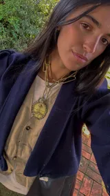 Colgante grande de estilo antiguo, con forma ovalada y borde festoneado, que representa el Escudo Nacional Argentino. Está unido a una cadena gruesa de eslabones rectangulares dorados.