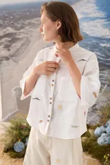 Camisa de lino blanca con cuello mao, mangas cortas y cierre frontal de botones. Presenta delicados bordados multicolores distribuidos por la prenda y bolsillos frontales en el pecho.