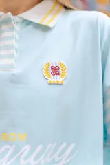 Polo de manga larga en color marrón con rayas horizontales azules en las mangas. Presenta cuello tipo polo en azul con detalles de rayas, cierre frontal parcial con botones y estampado frontal con la frase 'From Uruguay to the World'. Incluye un parche bordado en el pecho.