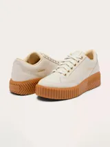 Zapatillas Sorel modelo Ona Ave Low, de diseño minimalista en color crema con suela de plataforma dentada en tono caramelo. Presenta cierre de cordones con ojales metálicos dorados y logo de la marca grabado en el lateral.