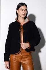 Chaqueta corta negra de paño con ribete de cuero color marrón en todo el contorno. Se ata con una tira de cuero en el centro y tiene dos bolsillos laterales.