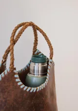 Bolso tipo matera de cuero con textura de reptil, color tostado, con manijas trenzadas y detalles de costura artesanal en los bordes.