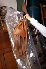 Cartera tipo morral de cuero color bordó, diseño tipo saco con cierre fruncido mediante cordones y flecos largos colgantes en la base. Cuenta con correa larga desmontable y logo grabado en la parte frontal.
