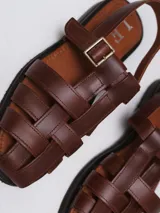 Sandalias tipo jaula de cuero marrón, con diseño de tiras entrecruzadas, cierre con hebilla metálica y suela plana.