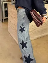Jean de tiro alto con pierna amplia de corte recto, confeccionado en denim de lavado claro. Presenta aplicaciones de estrellas en contraste con bordes decorados con tachas metálicas.