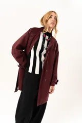 Trench de corte holgado color bordó, con cuello camisero, cierre frontal con botones y cinturón ajustable con hebilla. Presenta mangas largas con detalles de correas en los puños y bolsillos laterales.