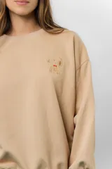 Buzo oversize de algodón frizado color beige, con cuello redondo, puños y cintura tejidos. Presenta un bordado pequeño de Snoopy en el frente.