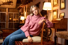 Blusa rosa de algodón con cuello alto y mangas largas abullonadas. Presenta detalles de cinta negra en el cuello y los puños, hombros con diseño princesa y cierre de lágrima con botón forrado en la parte posterior del cuello.