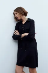 Vestido corto negro con cuello camisero, mangas largas, cierre frontal con cordones y bolsillos de parche en el pecho y las caderas.