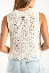 Top sin mangas de tejido de punto calado, color blanco, con cuello redondo acanalado y diseño de trenzas verticales. Presenta un ruedo con terminación festoneada.