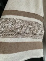 Saco tipo capa color beige, tejido en punto fino de lana con cierre de tancas de madera. Presenta cuello polo tejido y mangas anchas con detalles de trenzas y cintas tejidas.