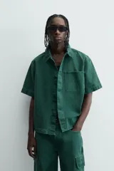 Camisa de denim de algodón color verde oscuro, de corte relajado (relaxed fit) y manga corta. Posee cuello solapa, bolsillo plastrón en el pecho con bordado a tono y cierre frontal de botones.