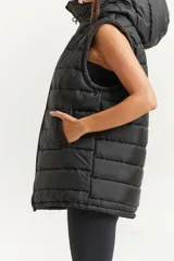 Chaleco negro oversize con capucha forrada en tela gamuzada, cierre metálico YKK, bolsillos laterales y tancas regulables en el ruedo.