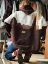 Campera tipo hoodie de textura tipo corderito, con diseño de paisaje montañoso en bloques de color marrón, blanco y beige. Presenta capucha y un parche de cuero con logo en la parte posterior.