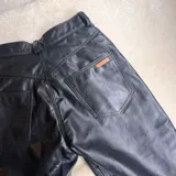 Pantalón de cuero vacuno negro, de corte recto y tiro medio. Presenta costuras horizontales a la altura de la rodilla y aberturas laterales en el ruedo. Incluye un botón de guampa irregular como detalle distintivo en la cintura.