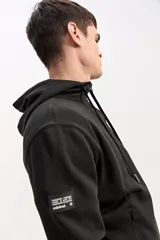 Campera de algodón gris melange con capucha, cierre frontal completo y bolsillos laterales. Cuenta con un parche rectangular negro con inscripciones en la manga y cordones ajustables para la capucha.