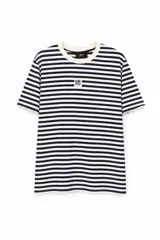 Remera de manga corta con diseño a rayas horizontales en color azul marino y beige. Presenta cuello redondo y un pequeño parche rectangular con estampado gráfico en el centro del pecho.
