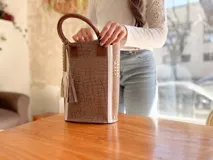 Bolso matero de cuero color taupe con textura croco y detalles en cuero color marrón. Tiene doble asa de mano y un dije de borla.