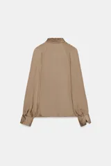 Blusa de tejido satinado color bordó, con cuello subido drapeado y manga larga con puños. Presenta detalle de pliegues frontales y cierre con botones laterales en el cuello.