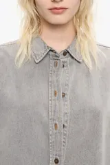 Camisa de manga larga color gris claro con efecto lavado, corte recto y botones frontales marrones. Presenta cuello camisero y pespuntes a contraste.