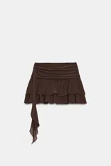 Falda pantalón de tiro bajo en color negro, con cintura drapeada y detalle de tira lateral colgante. Presenta un diseño con volantes en el bajo y cierre de cremallera metálica.