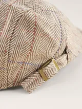 Boina estilo flat cap con diseño de espiga y cuadrillé en tonos beige, azul y rojo. Cuenta con una pequeña hebilla metálica ajustable en el lateral.